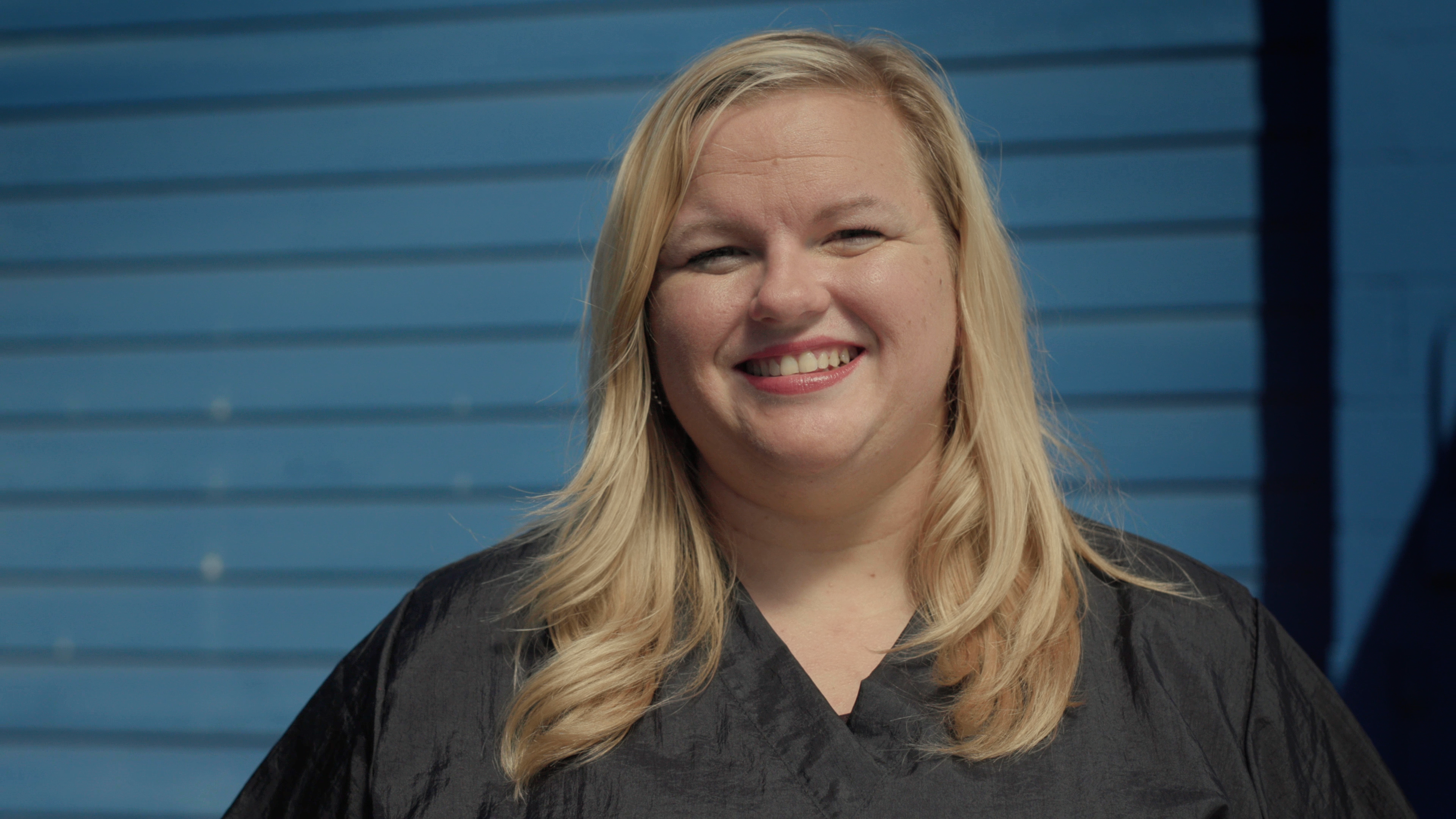 Close up of smiling female groomer in grooming smock with blue building out of focus in background