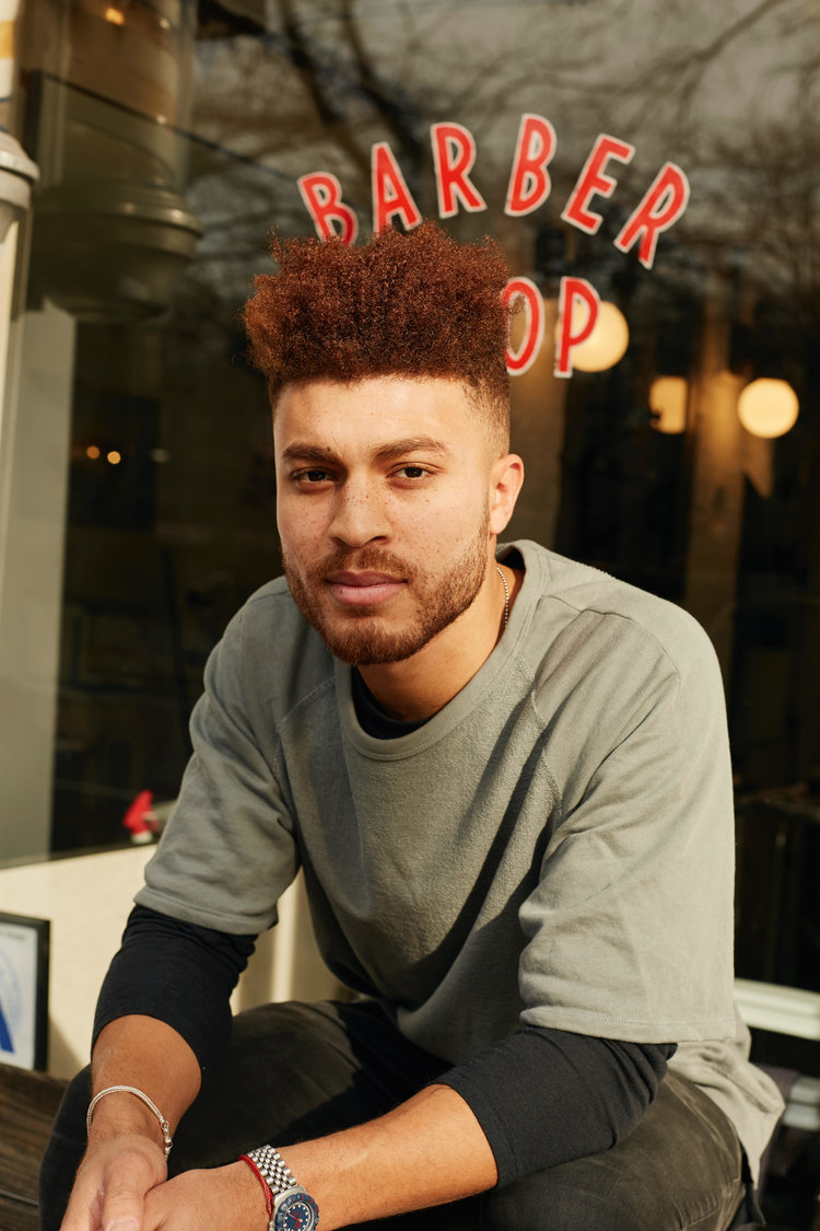 Julien Howard a.k.a. Velo Barber sitting in front of a barber shop window and looking directly into camera